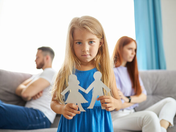 Ein Mädchen steht zwischen getrennten Eltern und hält Männlein und Weiblein aus Papier in seinen Händen
