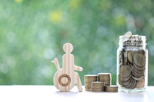 Ein Miniatur Holzrollstuhl steht neben einem Marmeladenglas voll Geld