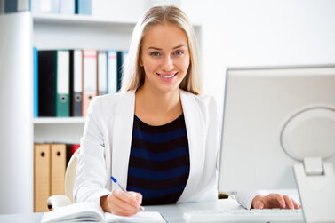 Eine junge Frau sitzt an einem Schreibtisch mit Computer und schreibt in ein Notizbuch