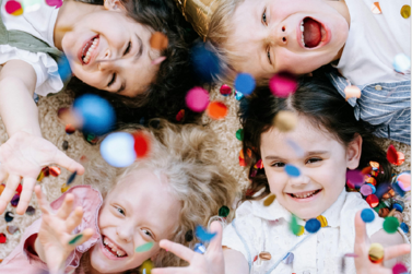 Vier Kinder liegen auf dem Boden. Man sieht die Gesichter der Kinder und wie diese Konfetti nach oben werfen.