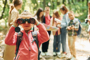 Mädchen mit Rucksack schaut durch ein Fernglas. Im Hintergrund eine Gruppe Jugendliche, die eine Karte studieren.