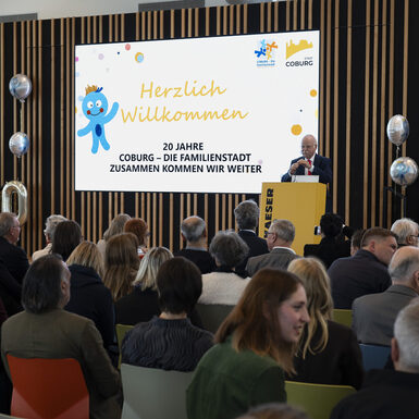 Publikum sitzt auf Stühlen vor der Bühne. Ein Mann steht am Rednerpult auf der Bühne.