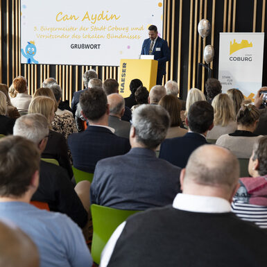 Publikum sitzt auf Stühlen vor der Bühne. Ein Mann steht am Rednerpult auf der Bühne.