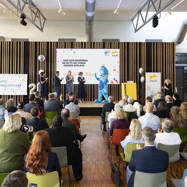 Publikum sitzend auf Stühlen vor der Bühne. Auf der Bühne stehen mehrere Personen und ein Maskottchen.