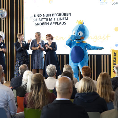 Publikum sitzend auf Stühlen vor der Bühne. Auf der Bühne stehen mehrere Personen und ein Maskottchen.