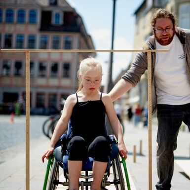 Ein Kind sitzt im Rollstuhl und testet eine Parcour aus. Ein Mann steht schräg dahinter und gibt Hilfestellung
