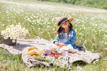 Ein kleines Mädchen sitzt auf einer Picknickdecke in einer großen Wiese. Sie trägt einen viel zu großen Hut, der ihr ins Gesicht rutscht.