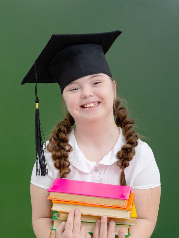 A teenager girl with Down syndrome wearing a graduate hat standing on the background of a green school board with a stack of books in her hands. Disabled girl with a smile looking at the camera