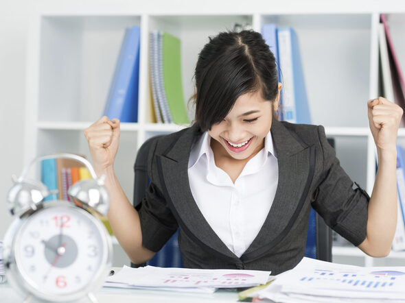 Junge Frau am Schreibtisch mit Unterlagen und Wecker. Sie streckt die Fäuste nach oben.