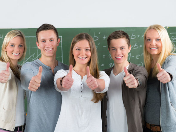 Fünf lächelnde Jugendliche stehen in einem Klassenraum vor der Tafel und zeigen