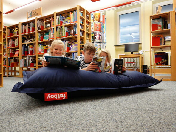 Zwei Jungen und ein Mädchen liegen mit Büchern in der Hand auf einem Sitzsack in der Kinder- und Jugendbücherei